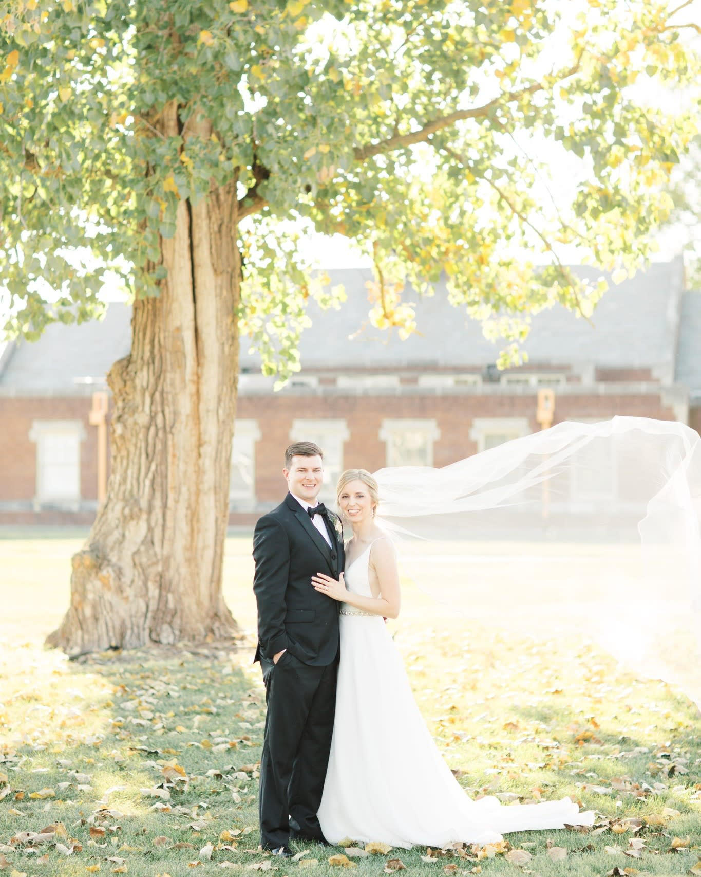 Bride and groom outdoors beneath a tree in autumn, with the historic brick venue behind them at 1899 Events